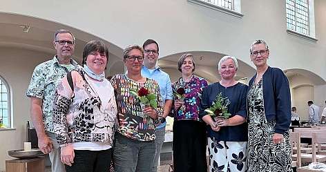 Eine Gruppe von sieben Personen posiert mit Blumen in einem hellen Raum.