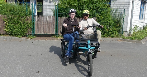 Zwei Personen sitzen auf einem Lastenrad in einer urbanen Umgebung.