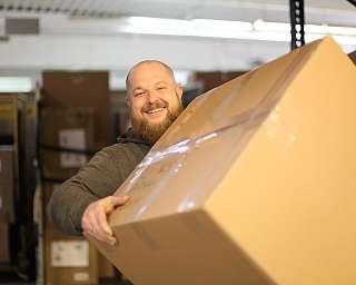 Ein lächelnder Mann hebt einen großen Karton in einem Lager.